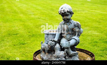 Statua di un ragazzino che tiene i suoi due cuccioli seduti su una base di legno con un prato verde nel terreno posteriore Foto Stock
