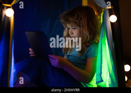 Un bambino piccolo usa un laptop di notte, un bambino siede nella tenda domestica davanti ad uno schermo del calcolatore nella stanza con il fulmine del neon, concetto del gadget Foto Stock