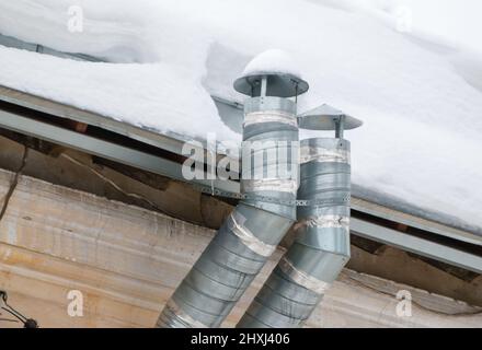 Due tubi di scarico in metallo sul tetto della casa. Sulla superficie c'è neve bianca. Sullo sfondo di un cielo grigio. Nuvoloso giorno d'inverno, luce tenue. Foto Stock