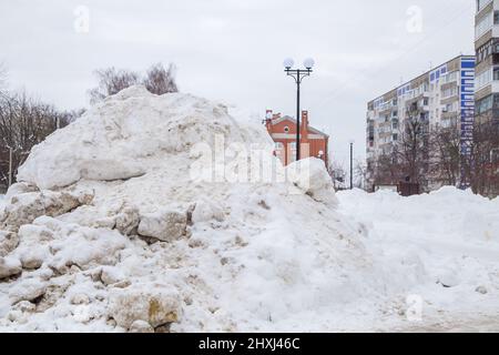 Una potente nevicata sulla strada contro lo sfondo di case e alberi della città. Sulla strada si trova neve sporca in cumuli alti. Paesaggio urbano invernale. Nuvoloso giorno d'inverno, luce tenue. Foto Stock