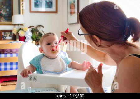 Bambino piccolo che è alimentato da sua nonna in un high-chair di plastica Foto Stock