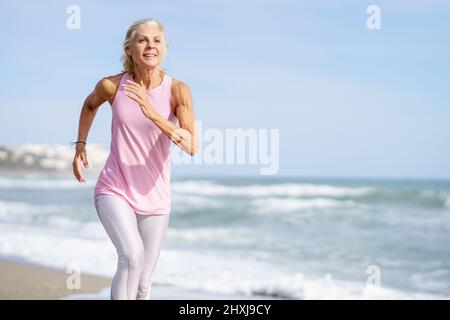 Donna matura che corre lungo la riva della spiaggia. Donna più anziana che fa sport per mantenere la misura Foto Stock