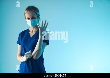 Giovane donna caucasica dottore che si mette su un guanto bianco sterile, si prepara per l'intervento chirurgico. Scatto medio in studio, sfondo blu. Foto di alta qualità Foto Stock