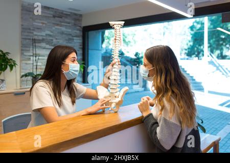 Fisioterapista che spiega la colonna vertebrale al paziente che indossa maschera chirurgica Foto Stock