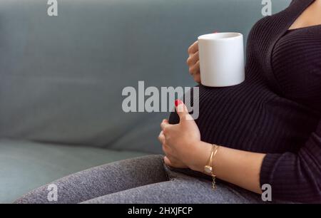 Mug bianco Mockup in mano di una donna incinta. Donna incinta che beve qualcosa da una tazza bianca. Donna incinta stanca che riposa. Mug mockup per designer Foto Stock