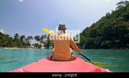 Giovane uomo con occhiali da sole e cappello ranghi rosa canoe di plastica lungo il mare contro verdi isole collinari con giungle selvatiche. Viaggiare in paesi tropicali. Il ragazzo forte sta navigando in kayak nell'oceano, vista posteriore. Foto Stock