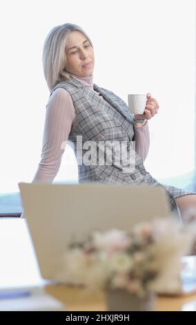 donna bionda che indossa abiti da ufficio seduti su un davanzale con una tazza di caffè che riposa. pausa caffè, giornata lavorativa dura. scatto verticale Foto Stock