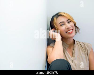 Felice donna asiatica ritratto con capelli corti in camicia beige senza maniche godere di ascoltare musica o canzone sulla radio con bianco wireless cuffie su wh Foto Stock