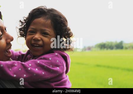 Java, Indonesia - 12 24 2020: Una ragazza che sembra felice quando tenuta e abbracciato dalla madre mentre nei campi di riso al mattino Foto Stock