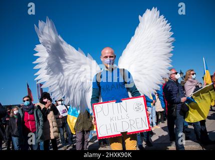 Amburgo, Germania. 13th Mar 2022. Un partecipante a una manifestazione contro il conflitto ucraino si trova su Jungfernstieg, indossando le ali di una colomba di pace e tenendo un segnale che dice "Putin uccide i bambini. Fermare la guerra ora'. Credit: Daniel Bockwoldt/dpa/Alamy Live News Foto Stock