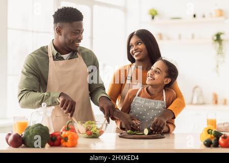 Genitori africani e figlia che fanno insalata insieme cucinare in cucina Foto Stock