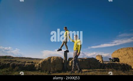 Uomo e donna in abbigliamento sportivo giallo verde. Una coppia incantevole di viaggiatori si abbraccia e bacia vicino alla pietra vecchia godendo il paesaggio delle Highland. Due viaggiatori camminano sullo sfondo di montagne innevate. Foto Stock