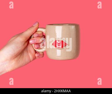 Tazza con tenuta a mano con immagine delle labbra su sfondo rosa corallo. Bacio. Foto Stock