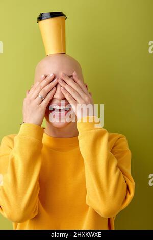 Divertente hairless femmina hanno divertente tenere una tazza di caffè sulla testa calva, godere la vita. Donna caucasica con malattia di allopecia che ride, vestita di grido casual Foto Stock