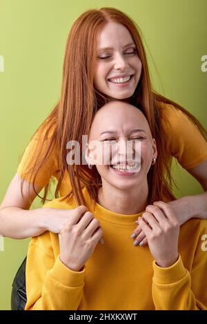 Due gioie giovani calva e rossa europee caucasiche donne abbracciano posa isolata su sfondo verde studio ritratto. Persone, concetto di emozioni umane Foto Stock