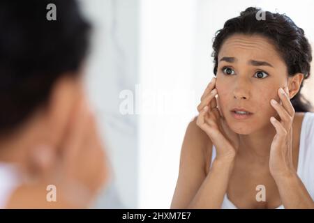 Concetto di pelle atopica. Giovane donna preoccupata che guarda lo specchio e tocca il volto Foto Stock