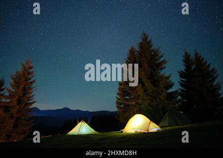 Luminose tende turistiche illuminate che brillano sul campeggio in montagne scure sotto il cielo notturno con stelle scintillanti. Concetto di stile di vita attivo. Foto Stock