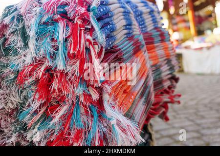 Coloratissimi pezzi di tessuto frangiati o tasseled in bazar donna produttore, pila di tessuto luminoso piegato primo piano sfondo. Vista laterale o profilo. Foto Stock