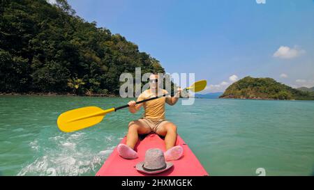 Giovane uomo con occhiali da sole e cappello ranghi rosa canoe di plastica lungo il mare contro verdi isole collinari con giungle selvatiche. Viaggiare in paesi tropicali. Il ragazzo forte sta navigando in kayak nell'oceano, vista frontale. Foto Stock