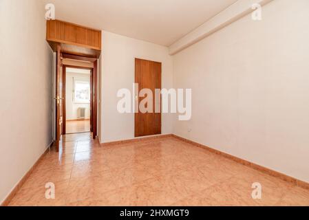 camera vuota con pavimento in gres arancio, porta in legno di mogano e pareti dipinte di bianco e maniglia in ottone su armadio e tronco Foto Stock