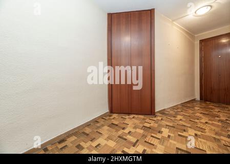 Sala vuota con pavimento in legno tipo sintasol, pareti dipinte di bianco, guardaroba da incasso con porte in legno e porta blindata coordinata Foto Stock