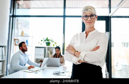Posso essere una mamma e un go-getter tutto in uno. Ritratto di una donna d'affari incinta in piedi in un ufficio con i suoi colleghi in background. Foto Stock