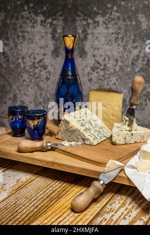 Ancora vita con formaggi assortiti, pecora stagionata, mucca di sale blu, brie e bottiglia di vetro blu con bicchieri, coltelli di formaggio e tavola di bambù Foto Stock