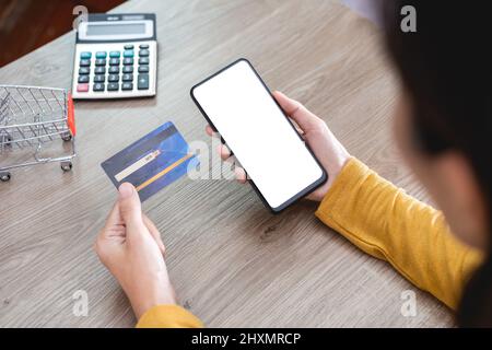 Le mani delle donne nella camicia gialla che tiene il telefono mobile con schermo vuoto e la carta di credito sulla scrivania dell'ufficio e usando il telefono mobile per lo shopping in linea, Intern Foto Stock