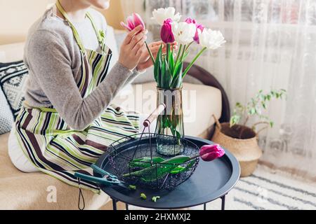 Donna mette bouquet di fiori tulipani in vaso con acqua a casa. Fiori freschi bianchi e viola raccolti nel cestino. Interni e decorazioni Foto Stock