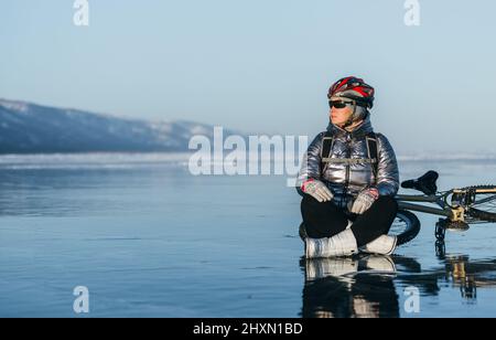 Donna seduta accanto alla sua moto su ghiaccio. Il ciclista della ragazza si fermò a riposare. Si siede al volante e gode della splendida vista del tramonto. Il viaggiatore è in bicicletta. La donna è in bicicletta sul ghiaccio. Foto Stock