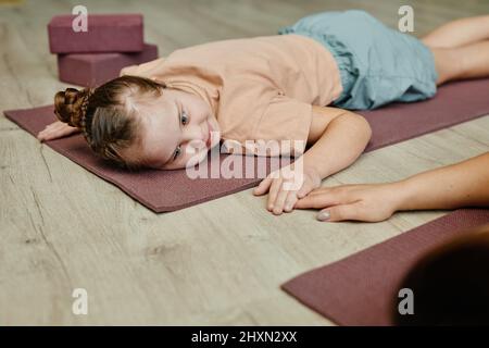 Ritratto di giovane ragazza con sindrome di Down che giace su uomo di esercizio a casa e tenendo le mani con la madre o l'istruttore aiutare Foto Stock