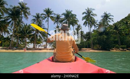 Giovane uomo con occhiali da sole e cappello ranghi rosa canoe di plastica lungo il mare contro verdi isole collinari con giungle selvatiche. Viaggiare in paesi tropicali. Il ragazzo forte sta navigando in kayak nell'oceano, vista posteriore. Foto Stock