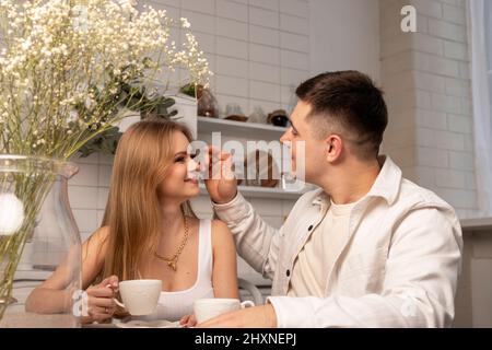 Giovane coppia seduta al tavolo da cucina, tazzine con tè o caffè, guardarsi e parlare. Felice moglie e marito. Foto di alta qualità Foto Stock