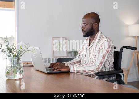 Giovane uomo d'affari serio o freelance in sedia a rotelle seduta da tavolo in soggiorno e guardando lo schermo del computer portatile durante la digitazione Foto Stock