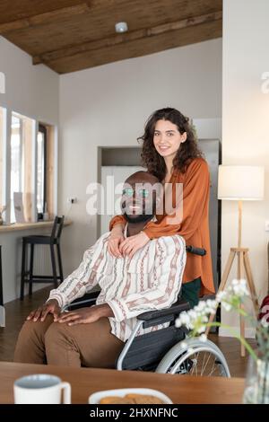 Felice giovane coppia interrazziale in casualwear guardando la macchina fotografica con sorrisi mentre si passa il tempo insieme a casa Foto Stock