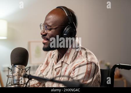Giovane calvo africano americano uomo in occhiali e cuffie cantando nel microfono mentre registra le sue canzoni in studio Foto Stock