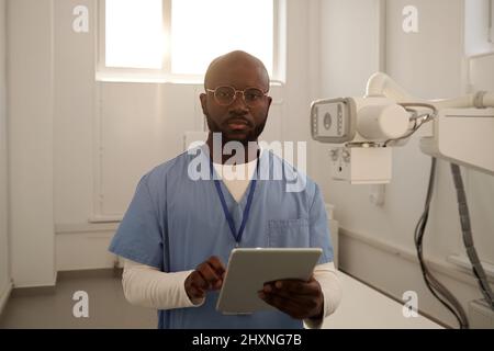 Giovane medico africano serio americano maschile in uniforme con tablet guardando la fotocamera mentre si consultano i pazienti online Foto Stock