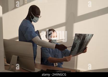 Giovane assistente afroamericano in maschera uniforme e protettiva che punta all'immagine a raggi X tenuta da radiologo femminile Foto Stock