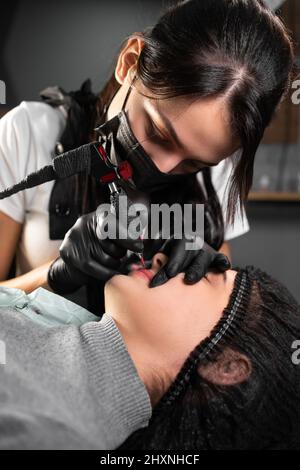 cliente femminile del salone di bellezza si trova sul divano. Estetista applica inchiostro permanente alle labbra. Procedura permanente di trucco del labbro. Foto Stock