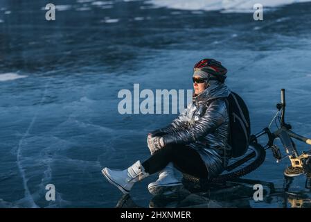 Donna seduta accanto alla sua moto su ghiaccio. Il ciclista della ragazza si fermò a riposare. Si siede al volante e gode della splendida vista del tramonto. Il viaggiatore è in bicicletta. La donna è in bicicletta sul ghiaccio. Foto Stock