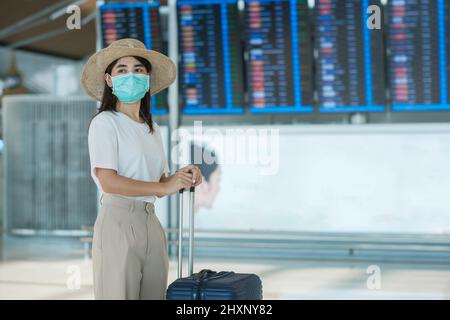 Giovane donna indossare maschera viso con bagaglio a piedi in aeroporto, protezione malattia di Coronavirus infezione, viaggiatore donna asiatica con cappello. Tempo di percorrenza Foto Stock