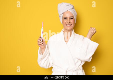 Donna felice in accappatoio tenuta gravidanza test in piedi isolato su sfondo giallo. Foto Stock