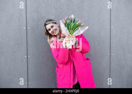 Ritratto di bella donna bionda stare vicino a parete di metallo con bouquet rosa di fiori omaggio celebrazione. Presente il giorno di marzo. Foto Stock