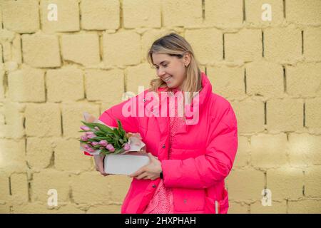 Felice bella donna in giacca, tenendo presente il pacchetto, tulipani fiori freschi, guardando il regalo di cura. Odore. Compleanno Foto Stock