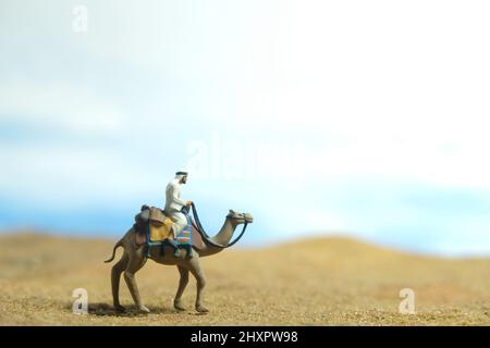 Fotografia di personaggi giocattolo in miniatura. Un uomo che indossa abiti bianchi kandura cavalcando cammello su un deserto in cielo luminoso. Foto immagine Foto Stock