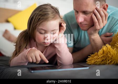 Padre e sua bambina con sindrome di Down sdraiati a letto e usando una tavoletta a casa. Foto Stock