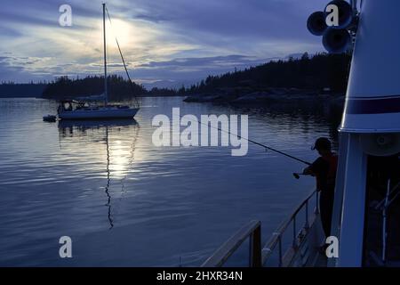 Un ragazzo che pesca con una canna che si staglia sul fianco di una grande barca al tramonto con una barca a vela delineata contro il suggestivo cielo blu scuro e bianco. Foto Stock