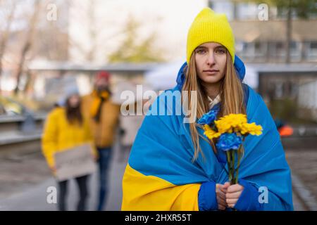 Protesta contro l'invasione russa dell'Ucraina. Giovane donna avvolta in bandiera Ucraina con fiori blu e gialli. Foto Stock