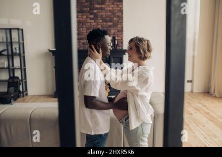 Il giovane uomo africano felice si alza, abbraccia la donna incinta caucasica e tocca il ventre con la mano. Concetto di matrimonio interrazziale. Foto Stock