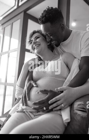 Il giovane uomo africano felice e la sua donna incinta caucasica si siedono insieme, si divertono e ridono con gioia. Concetto di matrimonio interrazziale. Imag bianco e nero Foto Stock